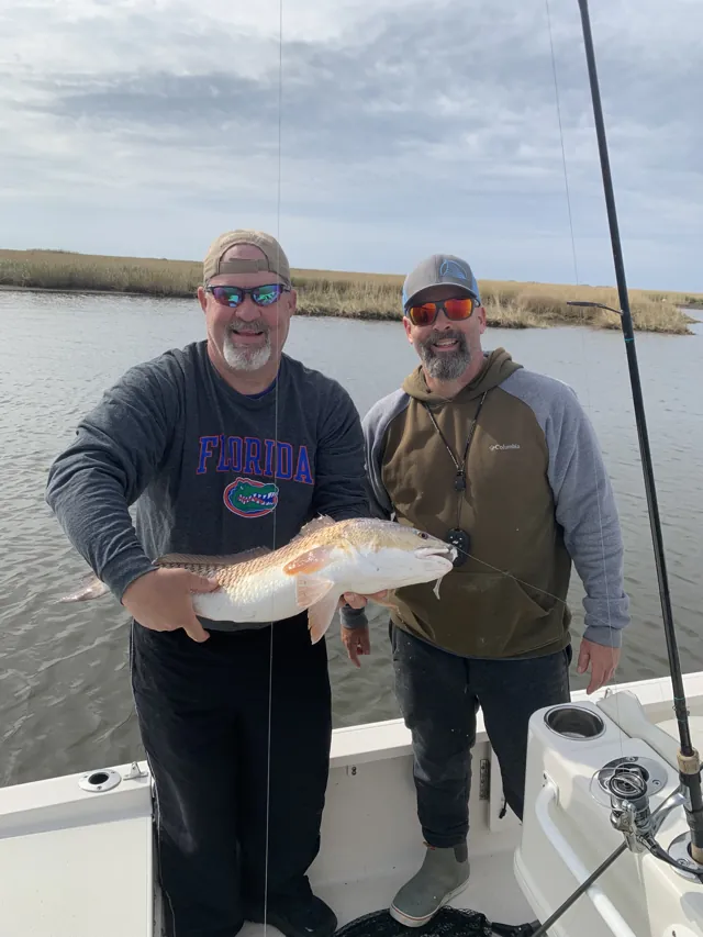Two anglers hold a redfish between them with marsh grass behind the boat.