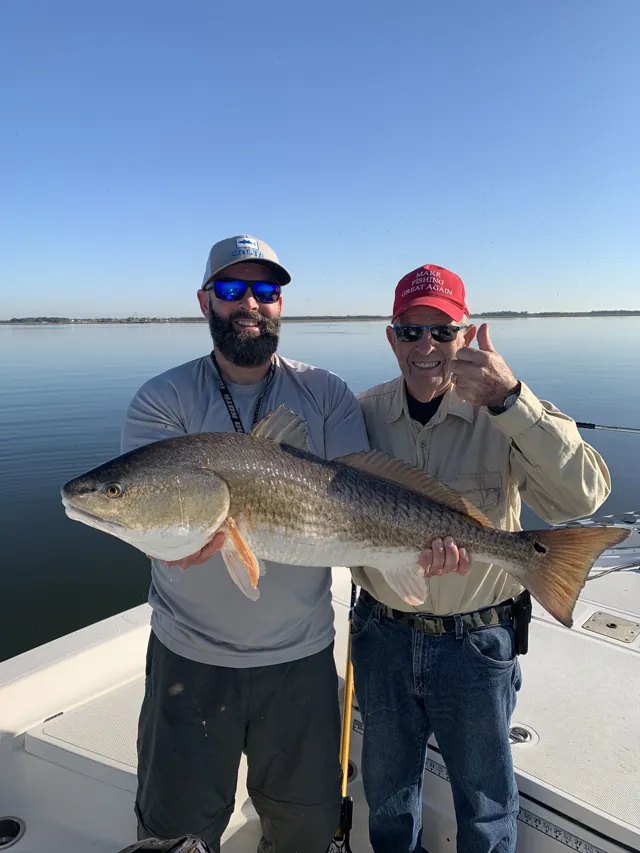 Two anglers on the deck hold up a long redfish, one in a red cap.