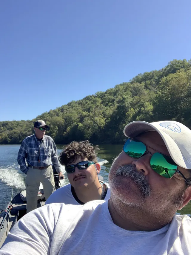 A selfie from the boat with two anglers behind, wooded hills along the river.