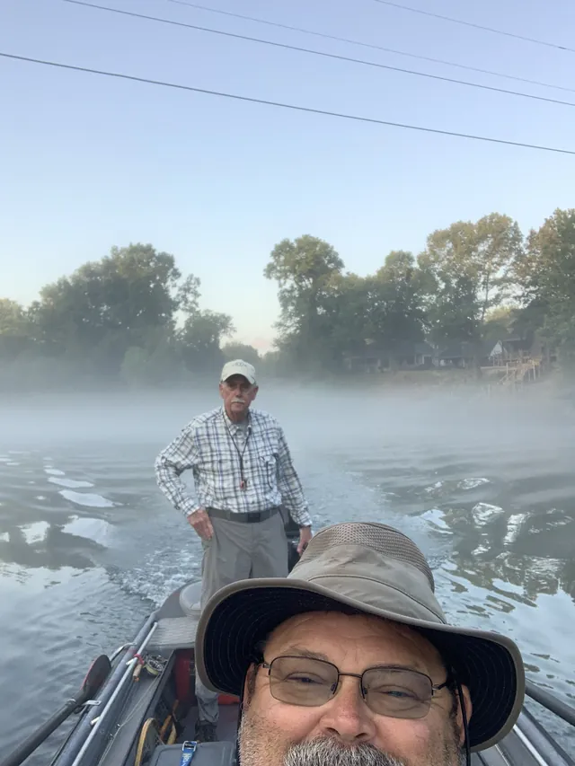 A selfie in a wide-brimmed hat, an angler standing in the boat on a misty river.