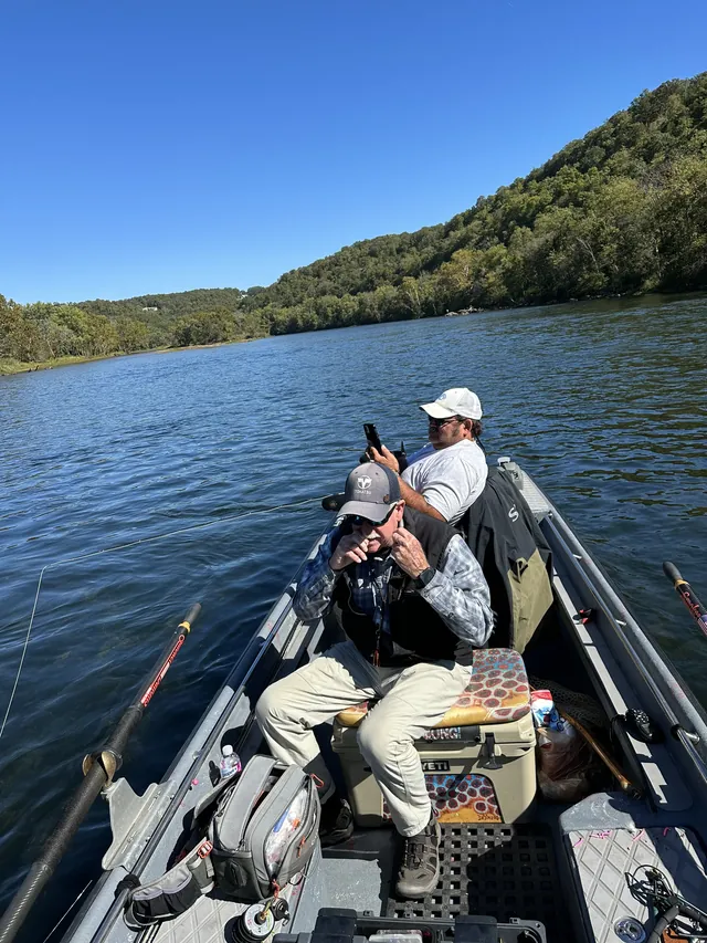 Two anglers in the bow of a river boat on wide green water, a wooded bluff behind.