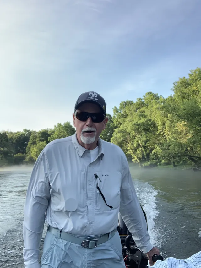 An angler in sunglasses stands at the tiller, river mist and trees behind.