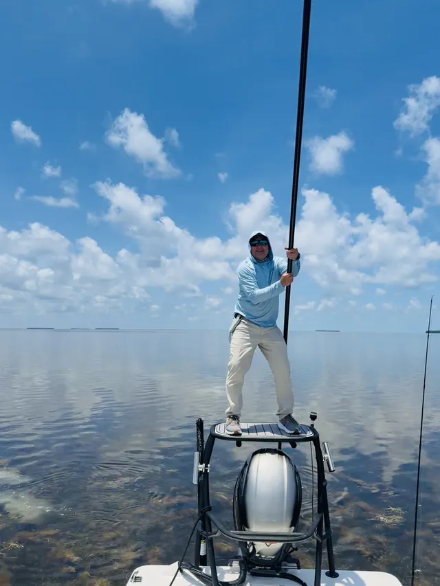 An angler stands on the poling platform of a skiff over flat, open water.